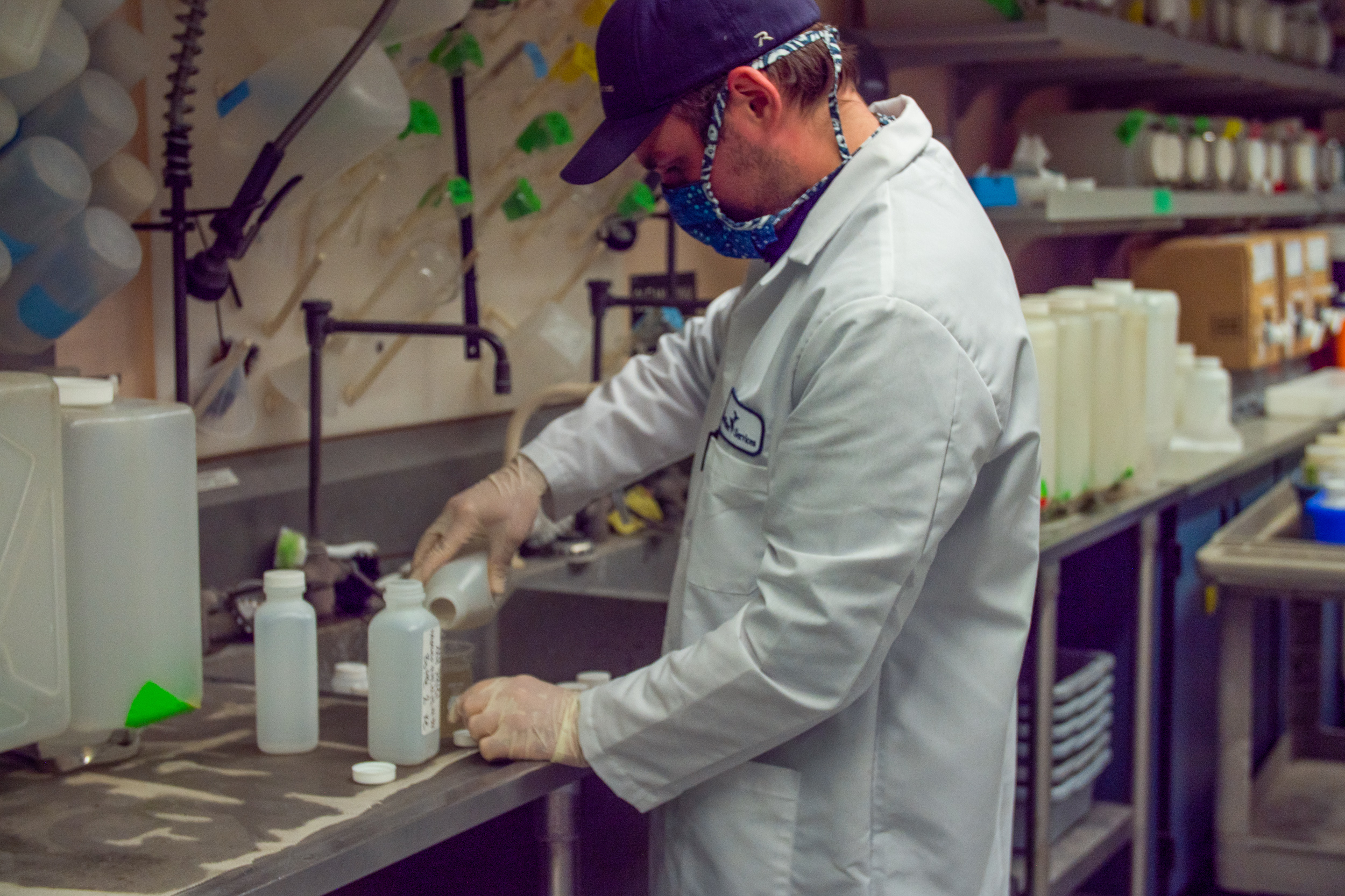 A Clean Water Services employee working in the lab. The employee wears a white lab coat, a protective mask, gloves, and a baseball cap. He pours liquid into a glass beaker