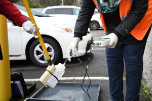 Un empleado de Clean Water Services usa tecnología para analizar el agua en una industria. El empleado lleva un chaleco naranja y guantes de goma.