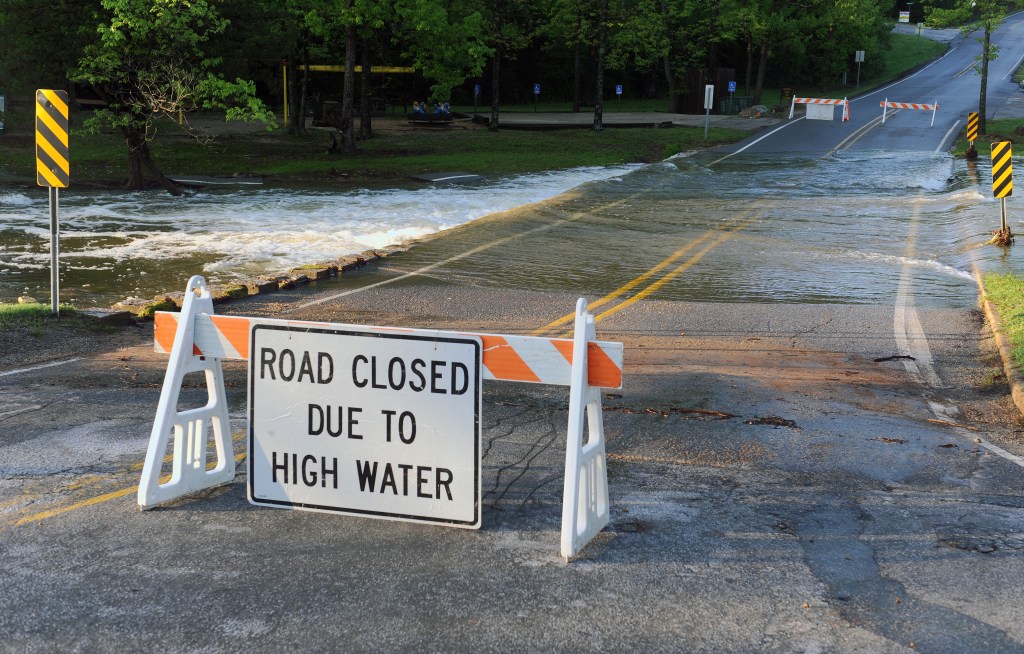 Un letrero de barricada que dice "Carretera cerrada debido a agua alta" se encuentra en una carretera frente a un área inundada de la carretera.