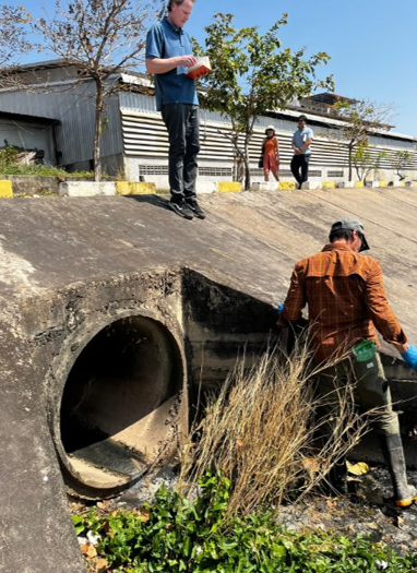 A person takes measurements next to the mouth of a large pipe coming out of a concrete structure. Another person stands above writing in a notebook. Two passersby, a few small trees, and a building are visible in the background. 