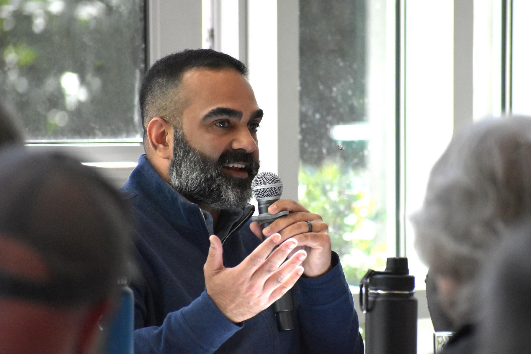 A person speaks into a microphone. The blurry heads of people listening are visible in the foreground.